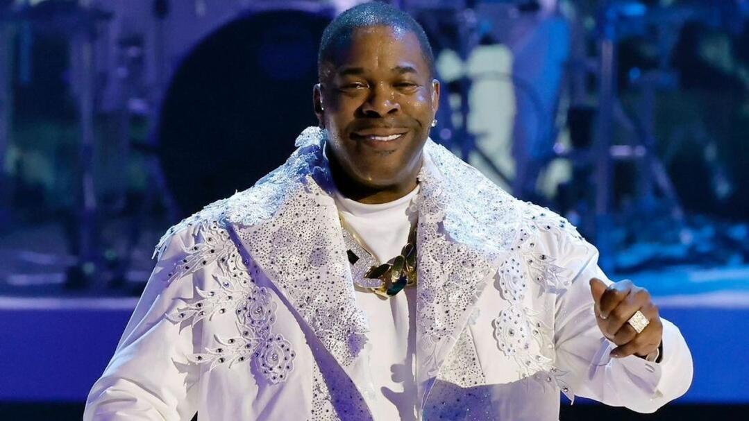 Busta Rhymes smiles in a white outfit with rhinestone bling, holding a microphone.