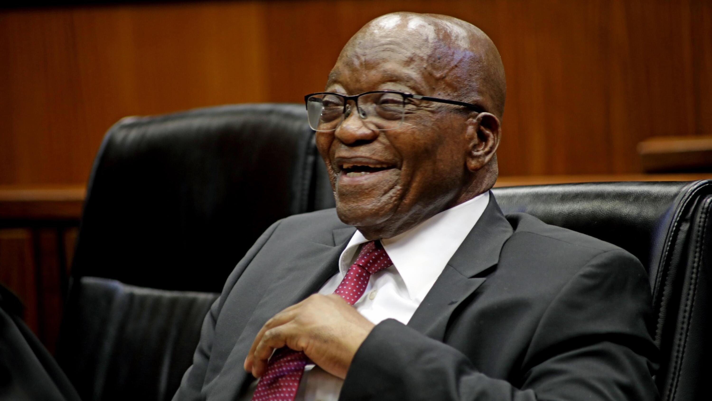 A smiling former president Jacob Zuma seated at a table.