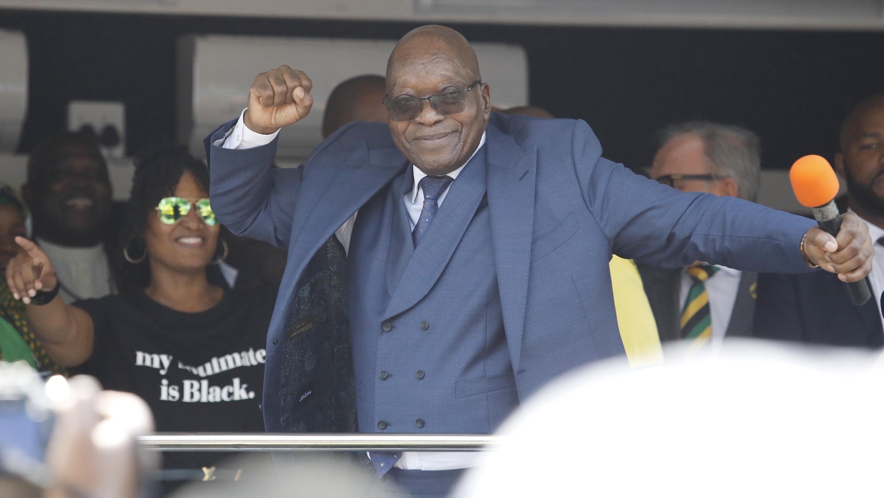 Former president Jacob Zuma dances on a platform with a mic in his hand. 