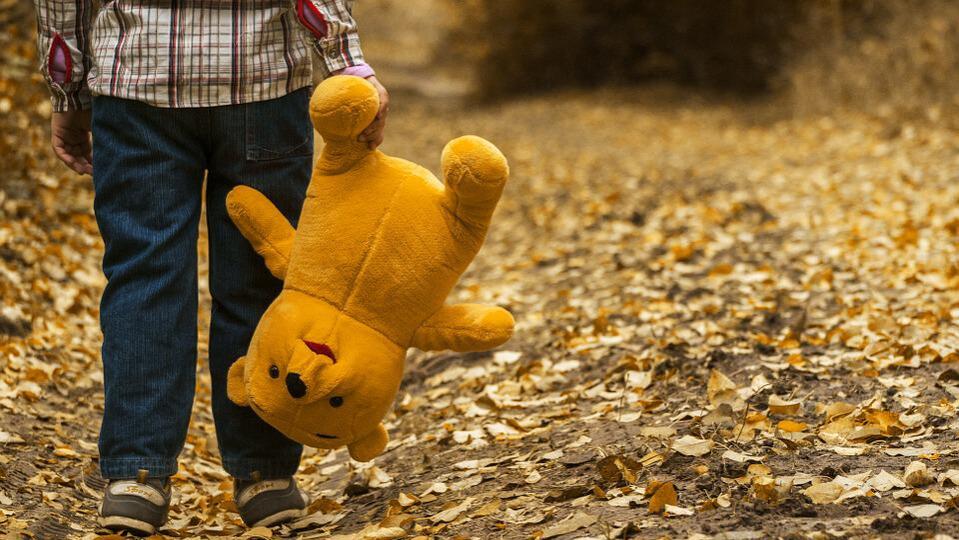 A boy walking with a Winnie the Pooh teddy bear hanging upside down. 