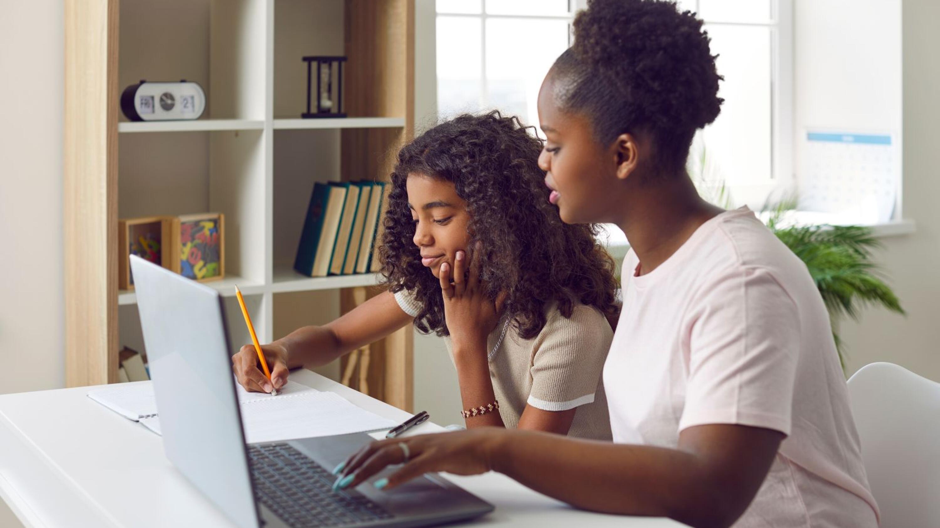 Family doing homework. Parent helping little child with homework. Young African American mother and her daughter sitting at desk at home, using laptop computer and writing essay in school notebook 